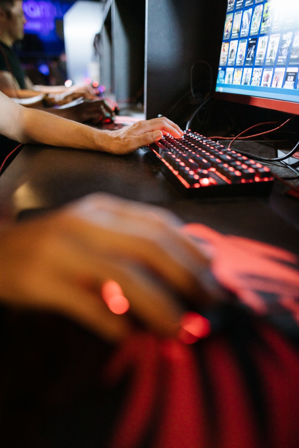 Hands on a backlit keyboard and mouse in a shared internet café with several workstations behind